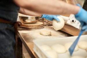 Elyas wearing blue gloves is handling freshly prepared dough in a professional kitchen, with a floured work surface and baking tools nearby.